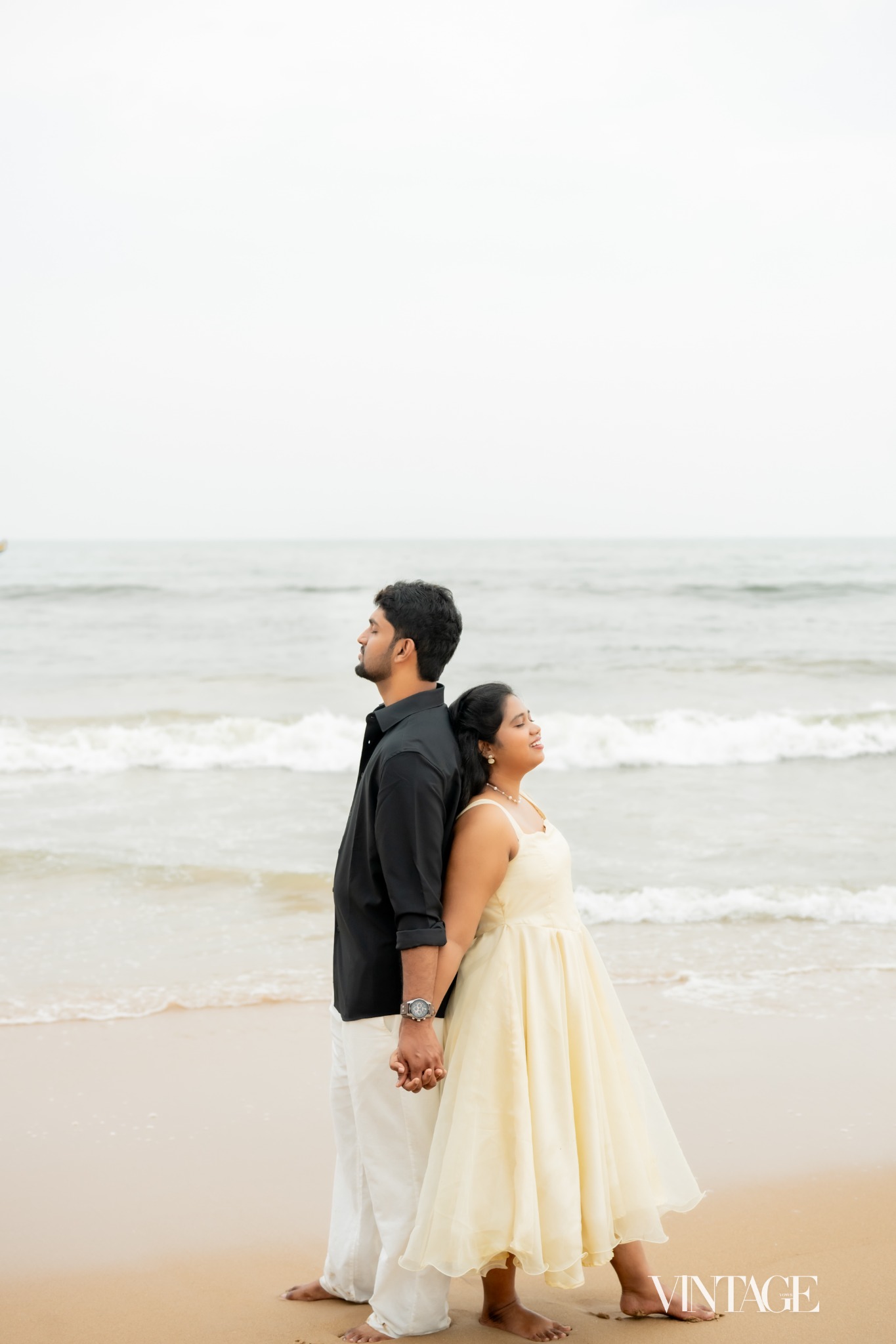 Ratan and Betsy standing back to back, holding hands beside the sea